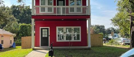 Une maison rouge à deux étages, dotée d’un balcon blanc, d’une boîte aux lettres et de deux voitures garées.