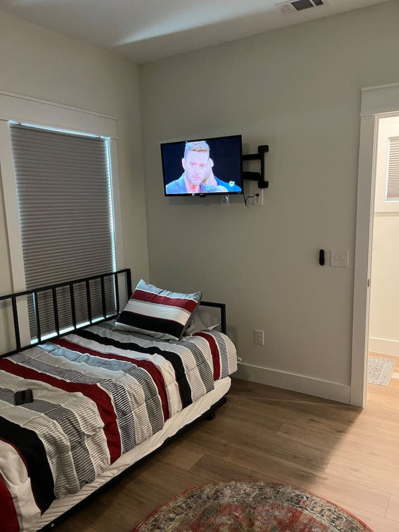 A bedroom with a bed, a television mounted on the wall, a window with blinds, and a door.