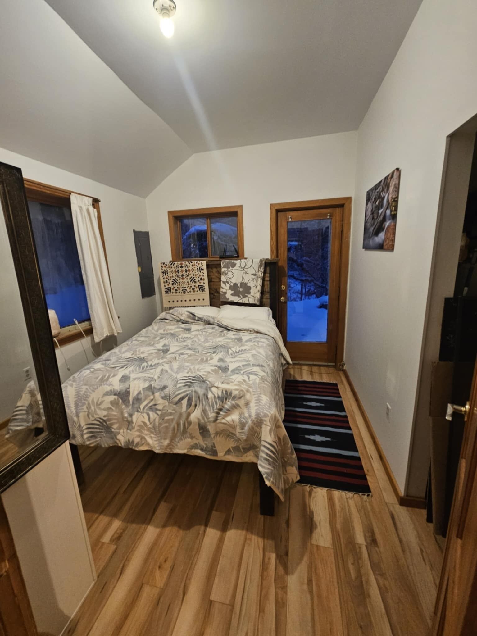A bedroom with a bed, wooden floor, two windows, a door, and a mirror.