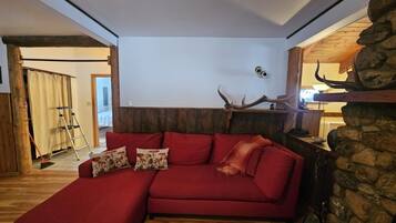 Un canapé d’angle rouge agrémenté de coussins à motifs floraux, un mur en pierre orné de bois de cerf et une échelle en bois dans une pièce dotée d’un plancher en bois et d’un plafond blanc.