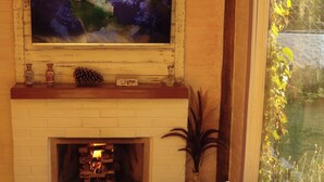 Un salon chaleureux avec une cheminée, un téléviseur fixé au-dessus et une fenêtre donnant sur un paysage verdoyant.