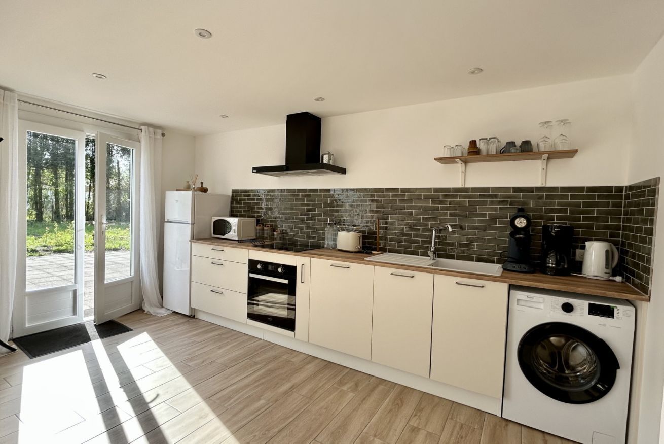 A modern kitchen with a built-in oven, microwave, and washing machine, wooden flooring, and a large window.