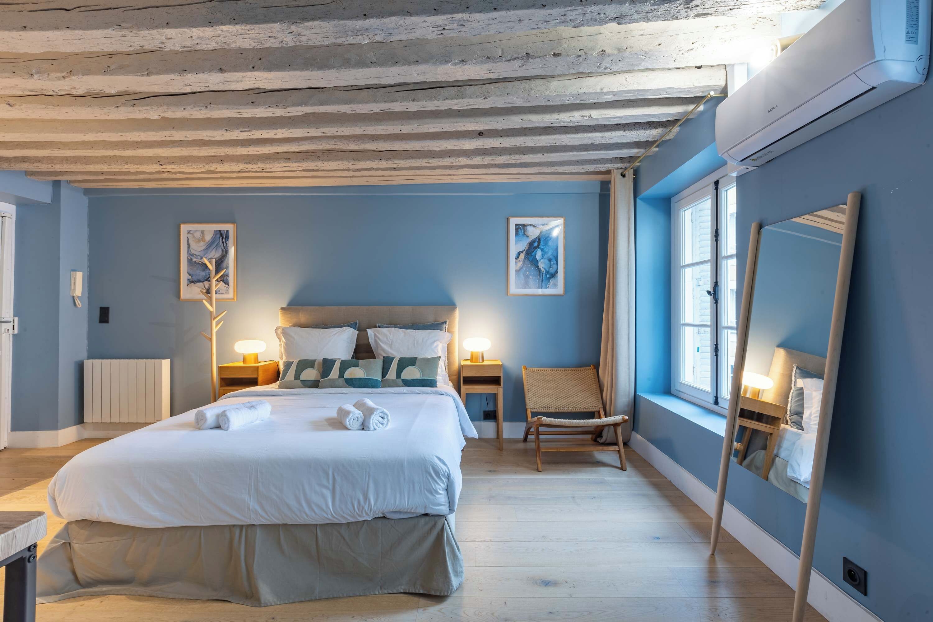 A bedroom with a large bed, bedside lamps, a chair, a window, and a mirror.