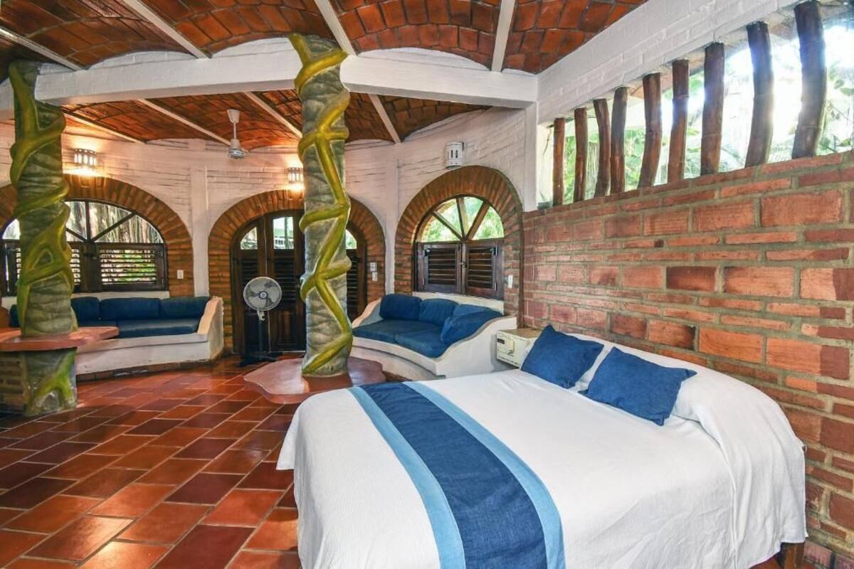 A room with a brick wall, a bed with blue pillows, a bench, and a fan.