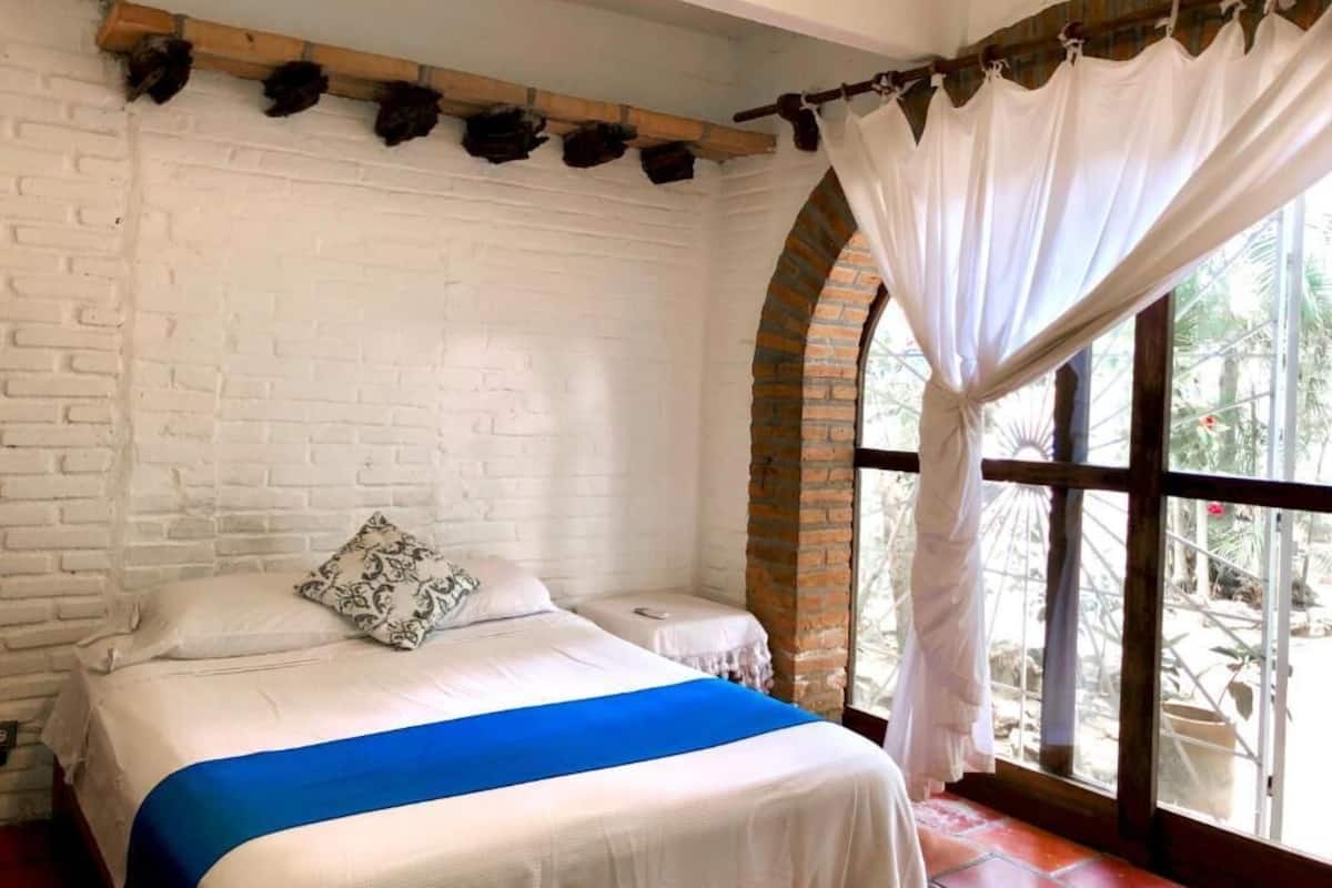 A room with a bed, white brick walls, a window with curtains, and a view of greenery outside.