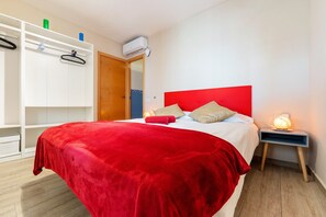 A bedroom with a red bedspread, a white wardrobe, a bedside lamp, and an air conditioning unit.