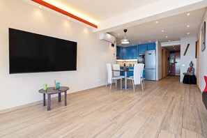 A modern living room with a flat-screen TV, a dining area with chairs, and a kitchen in the background.