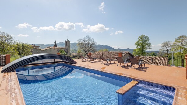 Ein Pool mit Fliesen, Liegestühlen und Blick auf Berge sowie eine Kirche im Hintergrund.