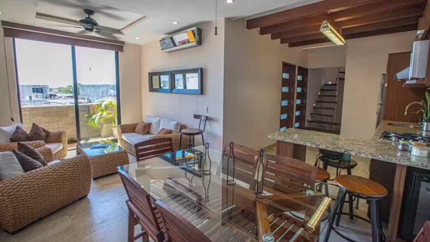 Un salon moderne avec une table basse en verre, des canapés en osier et un coin repas comprenant des chaises en bois et un bar.