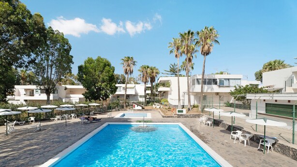 En swimmingpool med liggestole og parasoller, omgivet af palmer og moderne bygninger.