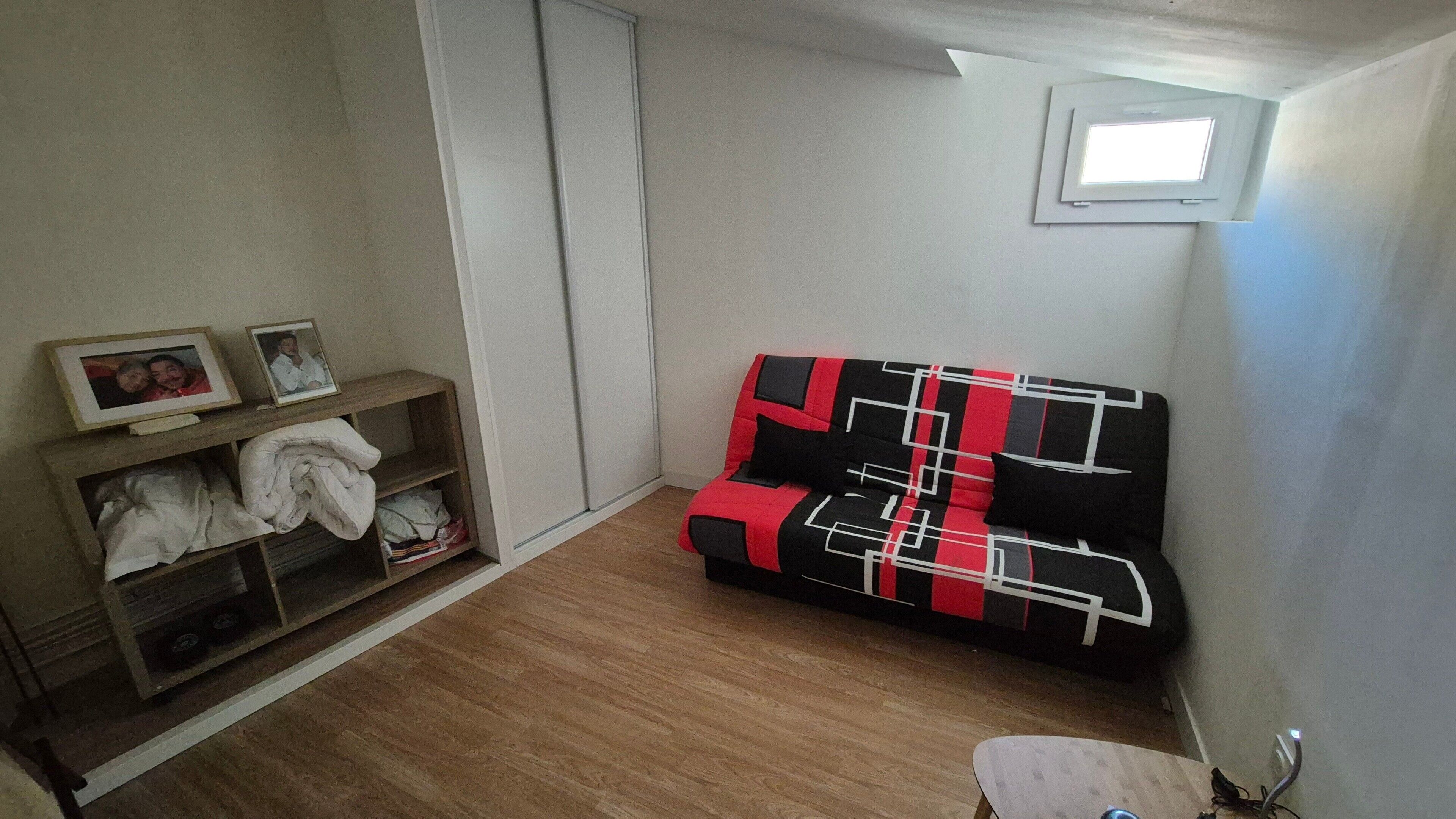 A small room with a wooden floor, a black and red sofa, a wooden shelf with folded clothes, and a small window.