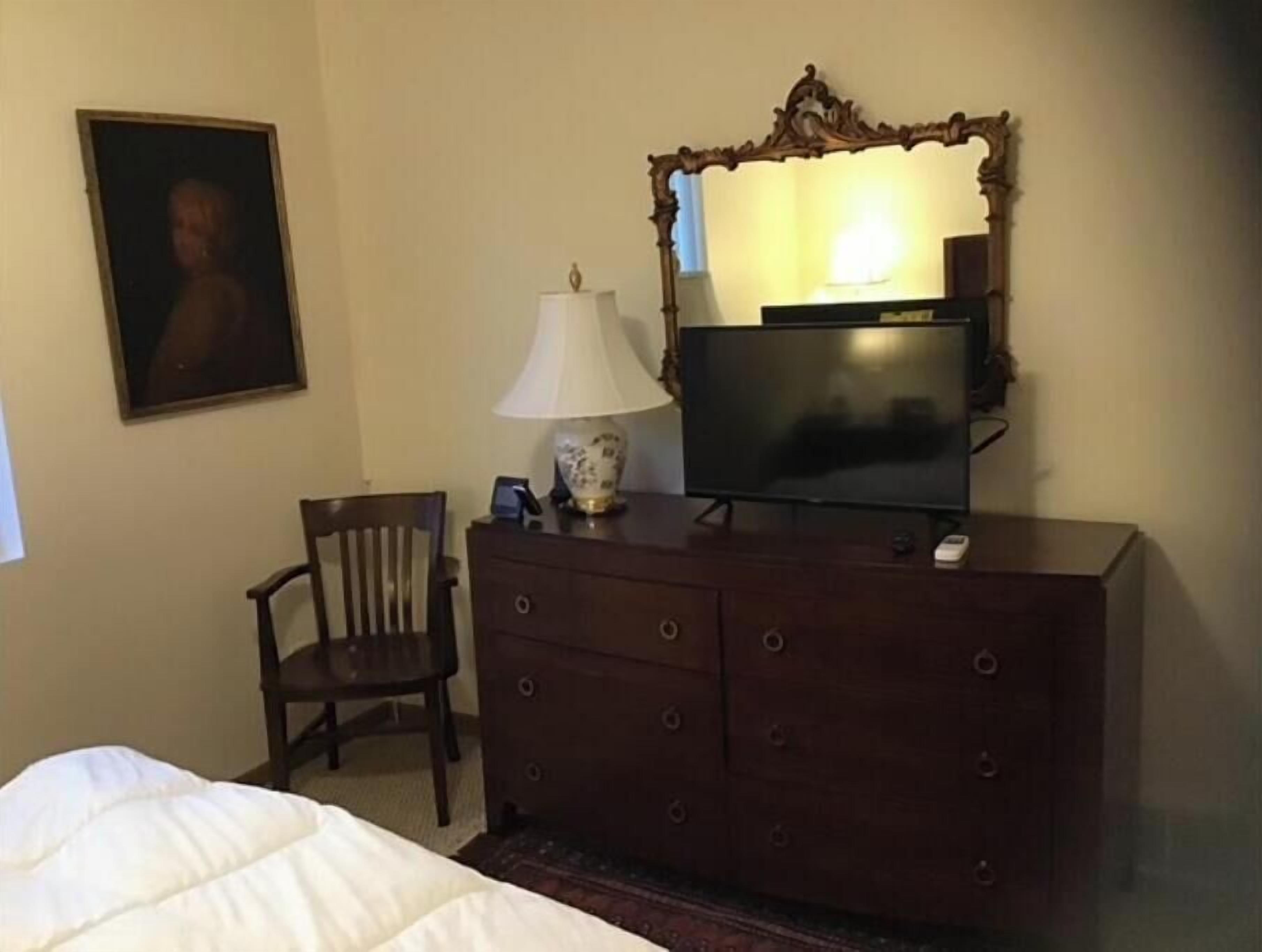 A hotel room with a bed, a wooden dresser, a television, a lamp, a chair, and a framed picture on the wall.