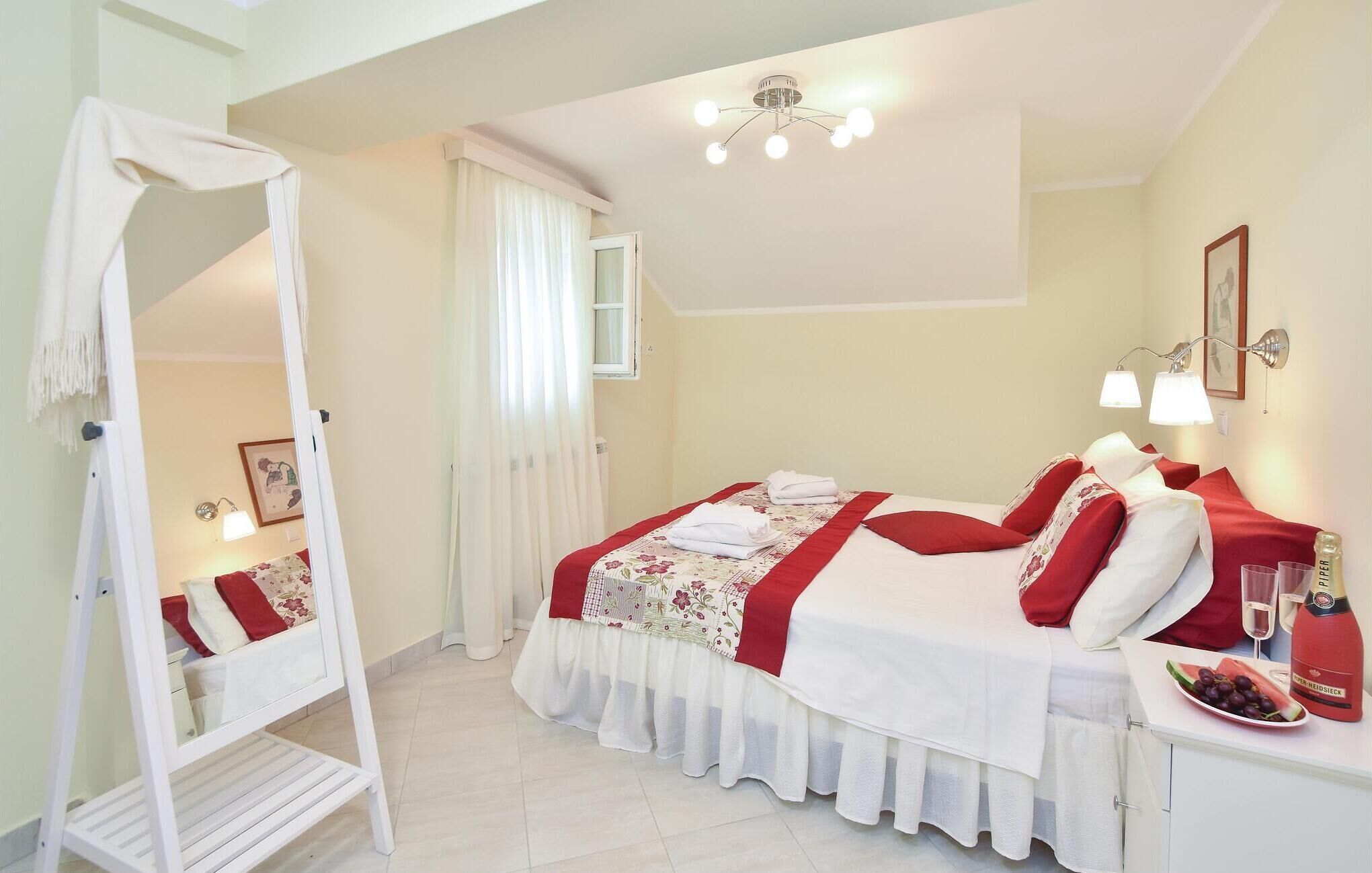 A bedroom with a bed, a bedside table with a bottle and glasses, a mirror, and a window with curtains.