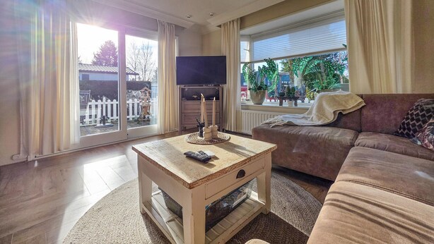 Un salon avec une table basse en bois, un canapé marron, un téléviseur et une vue sur un jardin grâce à de grandes fenêtres.