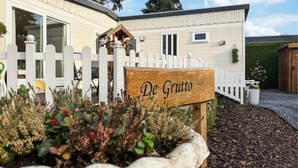 Une enseigne en bois portant l’inscription « De Grutto », placée devant une clôture blanche à piquets, avec une maison en arrière-plan.