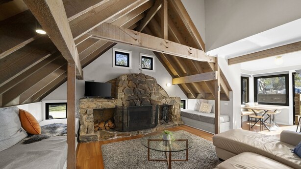 A cozy loft bedroom with a stone fireplace, wooden beams, and a large window. - Meltwater Chalet (Dinner Plain)