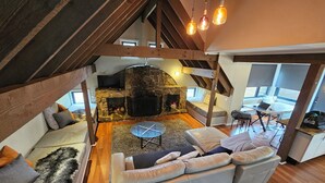 A loft-style living space with a stone fireplace, a sofa, a dining table, and a TV mounted on the wall. - Meltwater Chalet (Dinner Plain)