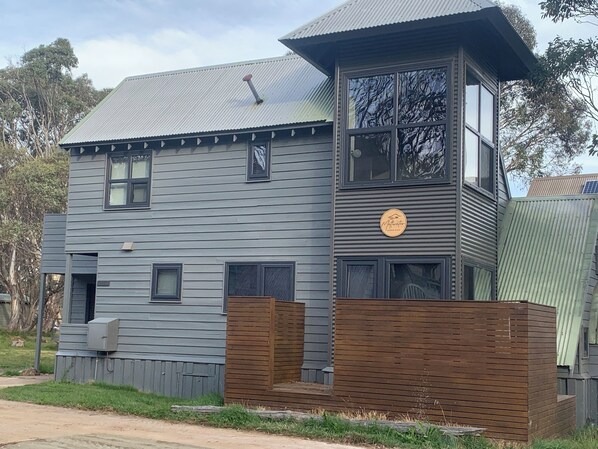 Un edificio de dos pisos con fachada gris, un porche delantero de madera y un ventanal grande en el nivel superior.