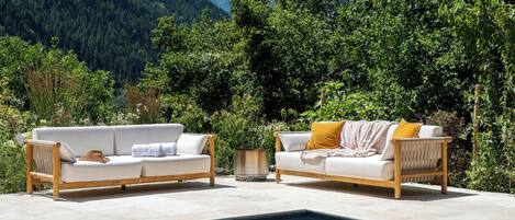 A poolside area with two white sofas and a small table, surrounded by lush greenery and mountains in the background.