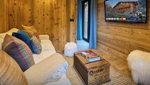 A cozy wooden interior with a sofa, cushions, a wooden coffee table, and a television displaying a mountain house. - Chalet Wonderland - Amazing chalet for 13 with summer pool - OVO Network (Séez)