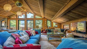 A spacious living room with a large sofa, a coffee table, and a television. The room has wooden beams on the ceiling and large windows. - Chalet Wonderland - Amazing chalet for 13 with summer pool - OVO Network (Séez)