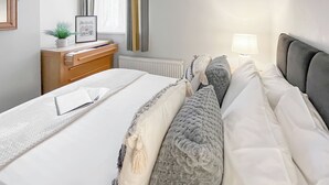 Ein ordentlich bezogenes Bett mit weißer und grauer Bettwäsche, ein Nachttisch mit Lampe und ein Fenster mit Vorhängen.
