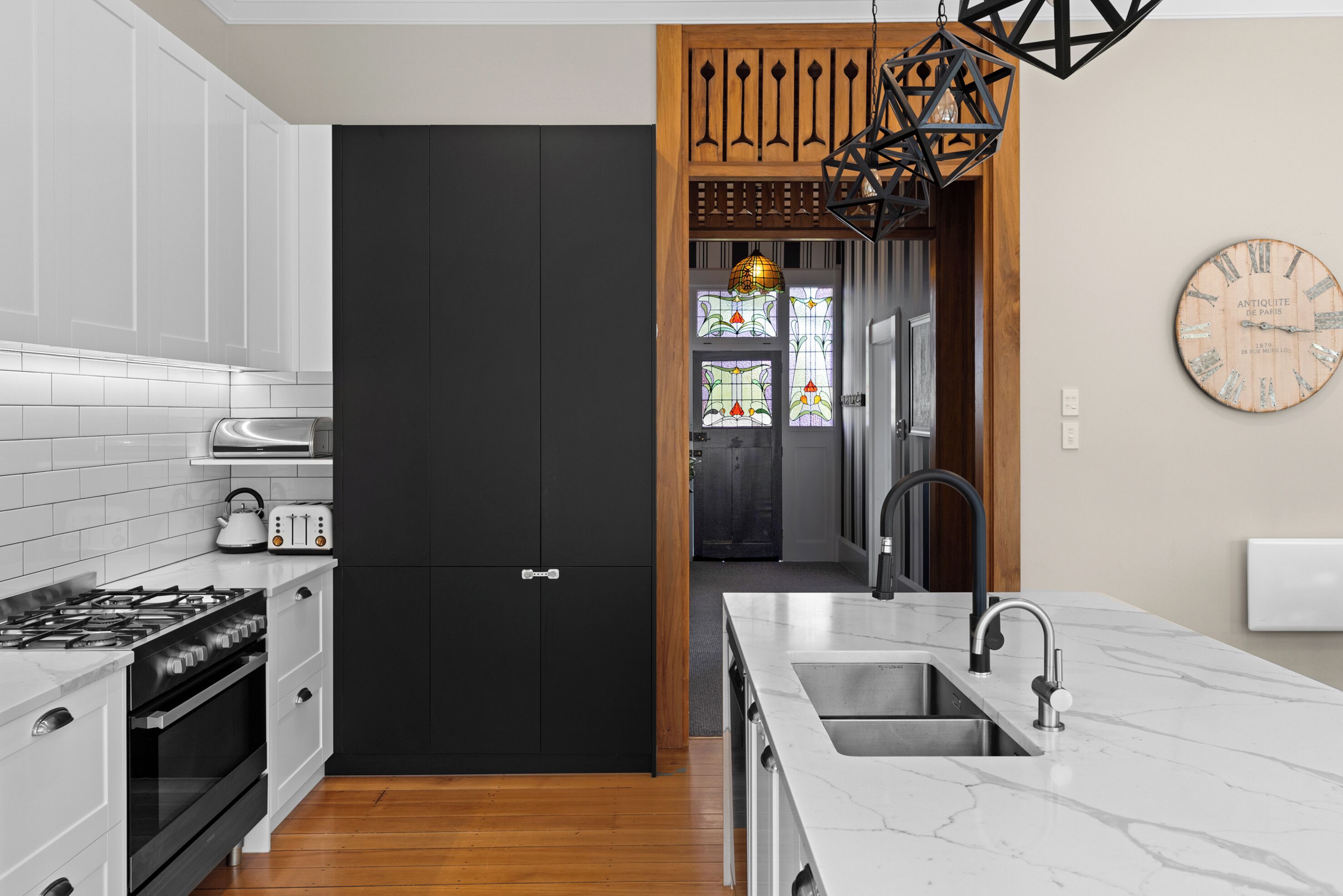 A modern kitchen with white cabinets, a black door, a wooden partition, a hanging light fixture, and a clock on the wall.