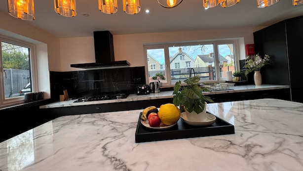 A modern kitchen with a marble countertop, a gas stove, and a black range hood. There are hanging pendant lights and a view of houses through large windows.