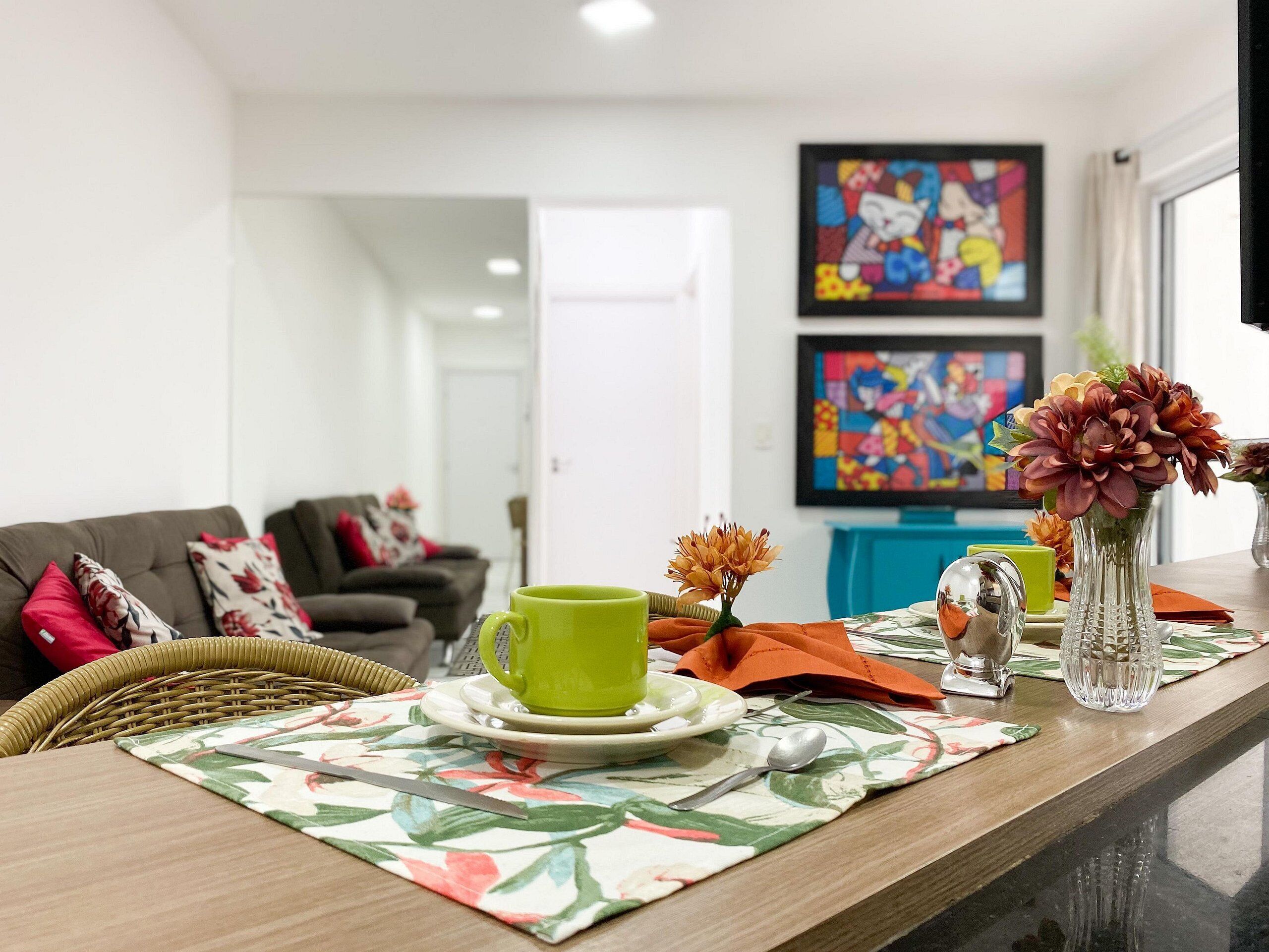 A dining area with a table set for two, featuring a floral table runner, a green cup, and a vase with flowers.