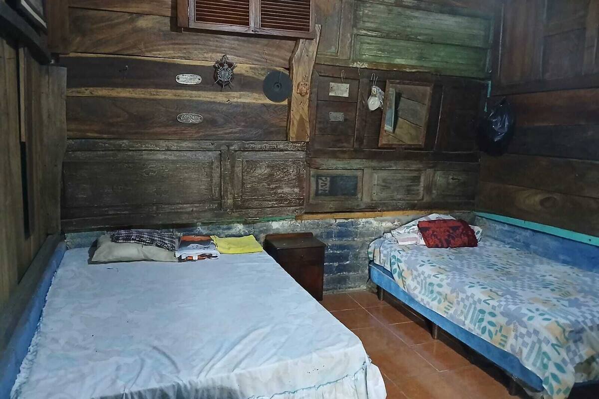 Quarto rústico de madeira com duas camas, uma com colchão branco e outra com padrão. Há almofadas e uma mesinha no quarto.