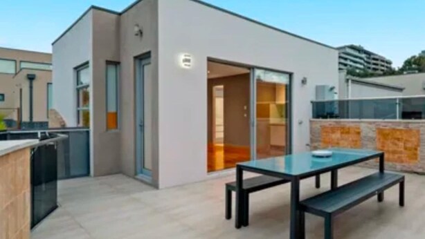 Een modern terras buiten met een glazen tafel en banken, een schuifpui die leidt naar een binnenruimte en een stenen muur.