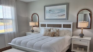 Un dormitorio con una cama grande, dos mesitas de noche, una silla de mimbre y un cuadro de una escena de playa.