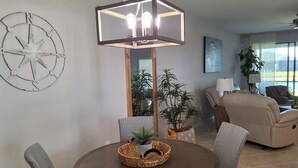 Un comedor con una mesa redonda, sillas de mimbre, una lámpara colgante, una brújula estelar montada en la pared y una planta en maceta.