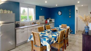 Een moderne keuken met een eethoek, met een grote tafel met een tafelkleed met geometrische patronen, rieten stoelen en een koelkast.