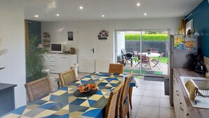 Een eethoek met een geruit tafel en rieten stoelen, een keuken met een koelkast en koffiezetapparaat, en een schuifpui naar een zitgedeelte buiten.