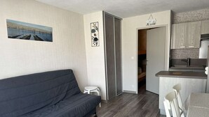 A compact living space with a grey sofa, a small white table, a kitchenette, and a door leading to a bedroom. - A practical one-bedroom flat with parking, close to the station and the beach (ARCACHON)