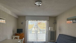 A room with a dining table, chairs, a sofa, a TV, and a window with a curtain. - A practical one-bedroom flat with parking, close to the station and the beach (ARCACHON)