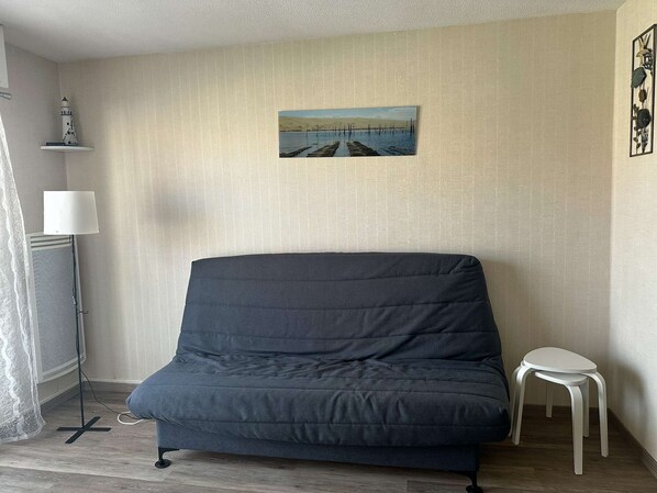 Une pièce avec un canapé bleu foncé, un petit tabouret blanc, une lampe sur pied et un tableau représentant un paysage côtier.