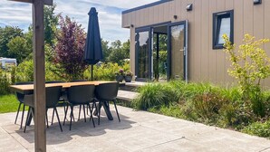 An outdoor patio with a wooden table and chairs, surrounded by greenery and a modern building with large windows. - 3 bedroom lovely (sta)caravan in Grou (Grou)