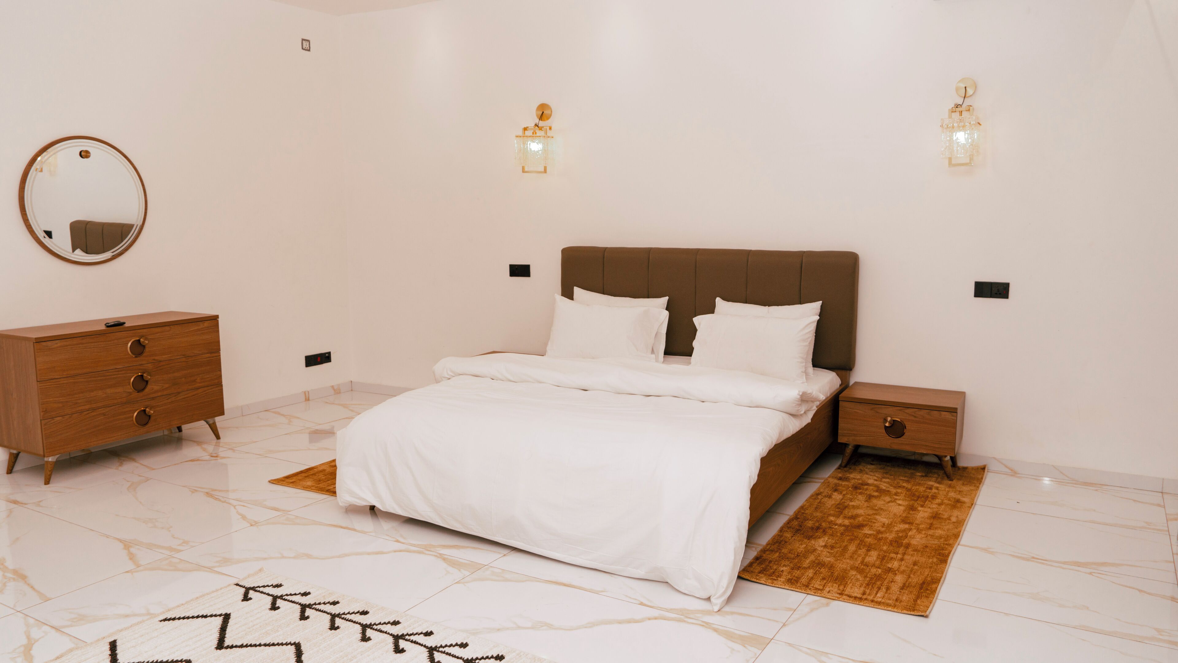 A bedroom with a bed, a dresser, a nightstand, a mirror, and wall sconces.