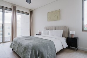 Relax in this serene bedroom with a large bed, nightstand, and wall-mounted artwork, featuring a view of the outside through large windows.