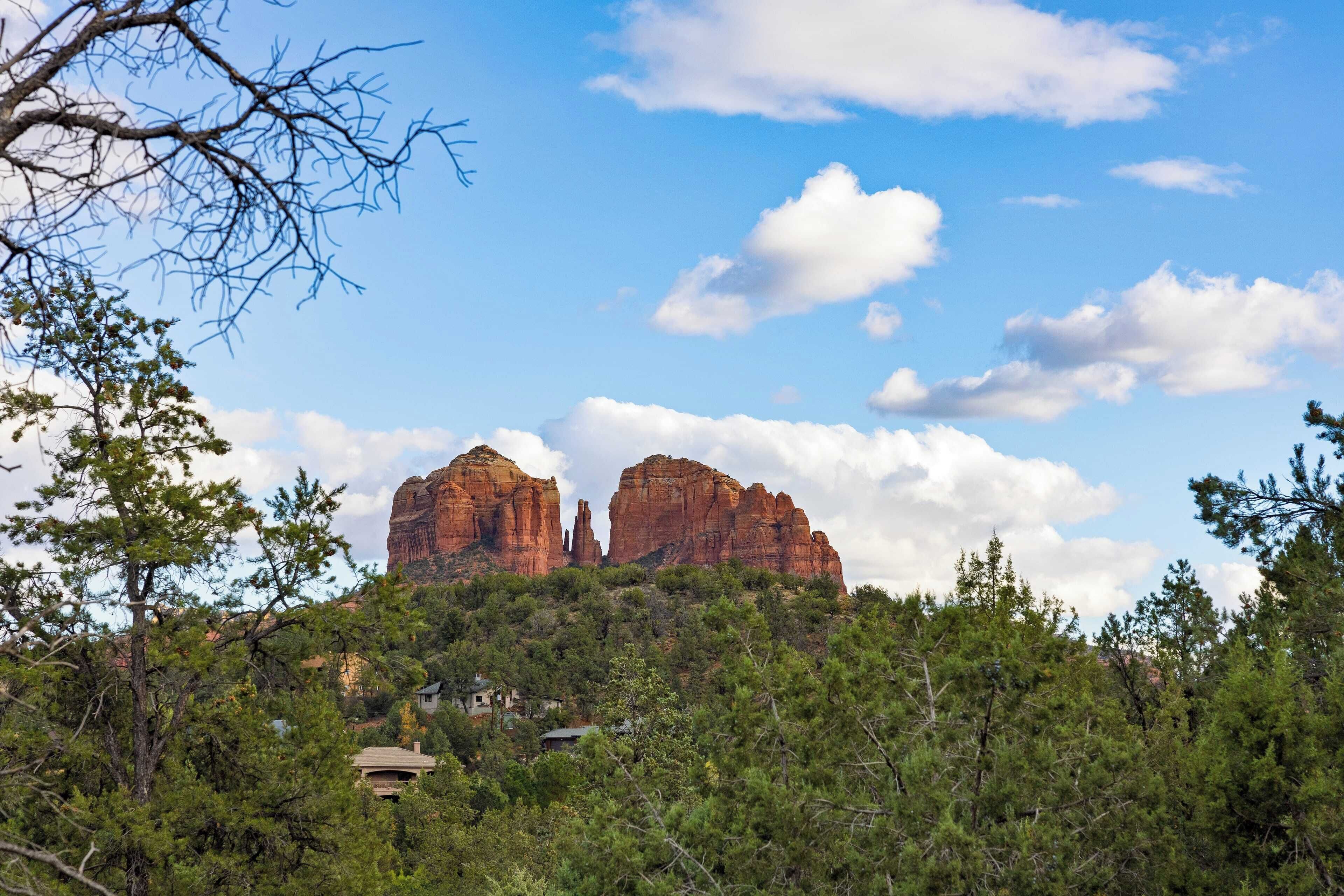 A mountain landscape with prominent red rock formations, surrounded by dense green forest and a few buildings at the base.