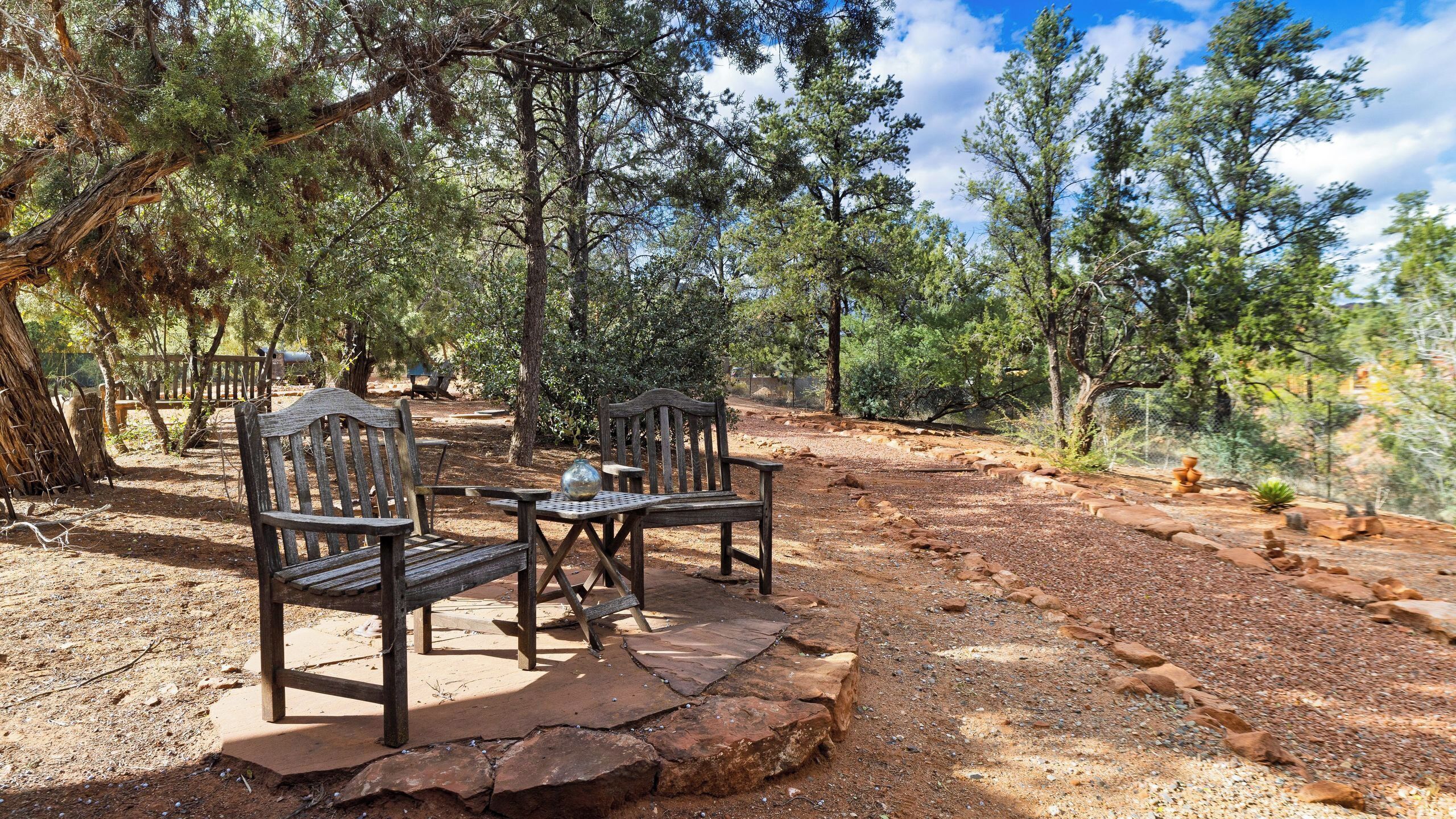 An outdoor seating area with wooden benches and a table, surrounded by trees and rocky terrain.