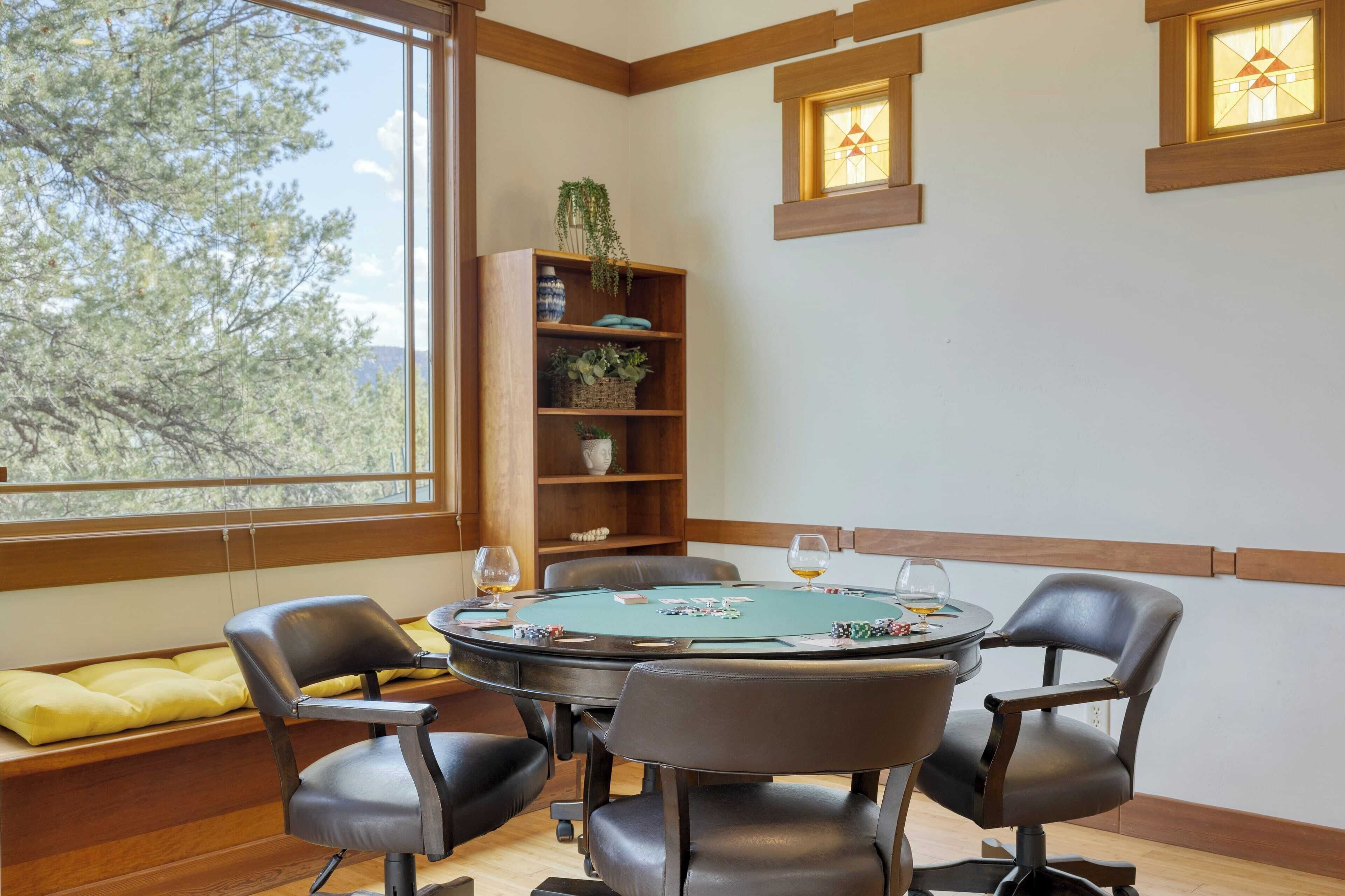 Une pièce comprenant une table de poker, quatre chaises, une bibliothèque et une fenêtre donnant sur des arbres.