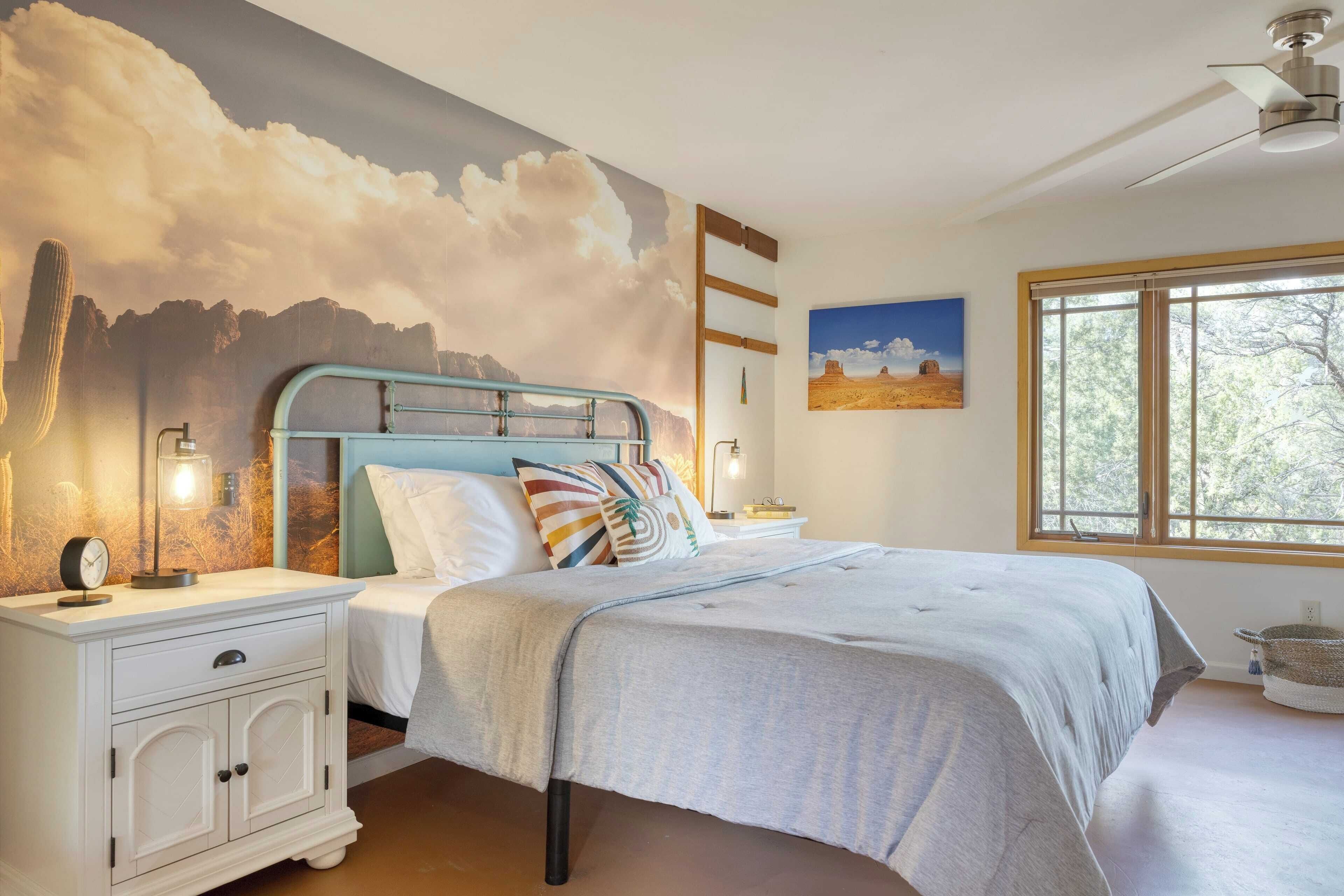 Une chambre à coucher comprenant un lit, des tables de chevet, une fresque murale, une fenêtre et un ventilateur de plafond.