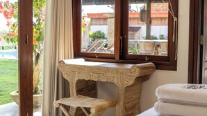 A room with a wooden vanity, a small stool, a window with a view of the outside, and a towel on the bed. - Tropical Villa Asmara (Ungasan)