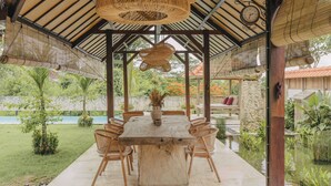 An outdoor covered dining area with a wooden table and chairs, surrounded by lush greenery and a view of a swimming pool. - Tropical Villa Asmara (Ungasan)