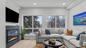 A modern living room with a fireplace, large windows, a grey sofa, and a painting on the wall. - NEW Private Hot Tub ~ Upscale Mountain Home (Canmore)