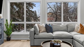 A modern living room with a grey sectional sofa, a wooden coffee table, and a large window offering a view of snow-covered trees and buildings. - NEW Private Hot Tub ~ Upscale Mountain Home (Canmore)