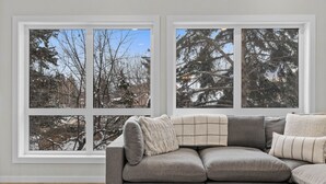 Ein Zimmer mit einem grauen Ecksofa, einem weißen Fellüberwurf und einem großen Fenster mit Blick auf schneebedeckte Bäume.